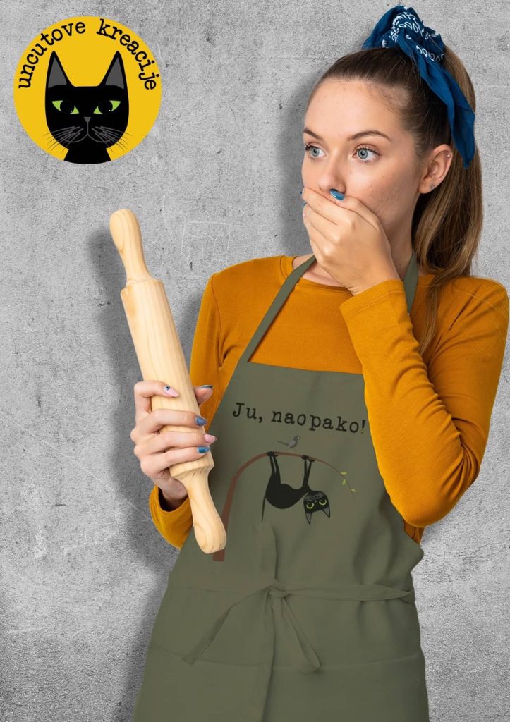 Fotografija prikazuje devojku koja nosi maslinastu kecelju sa ilustracijom crne mačke koja visi naopako na grani i natpisom „Ju, naopako!“ Devojka drži dlan preko usta, a u drugoj ruci joj je oklagija